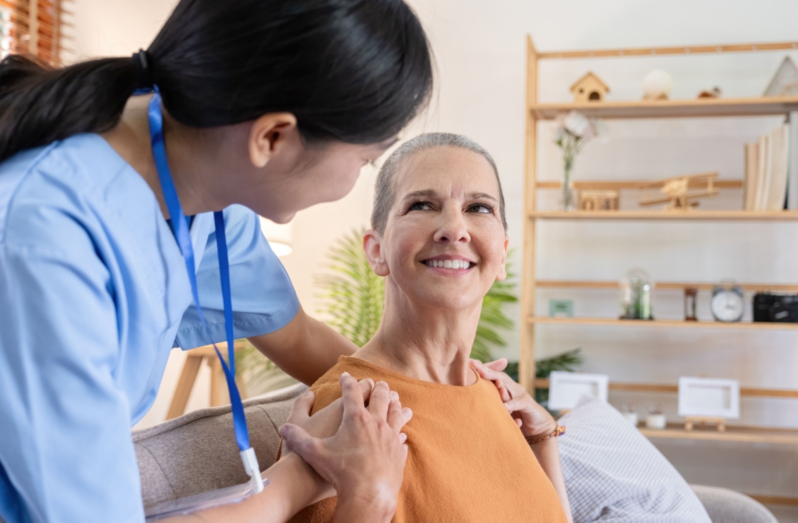 Caregiver in blue scrubs assists a smiling older woman seated on a sofa.