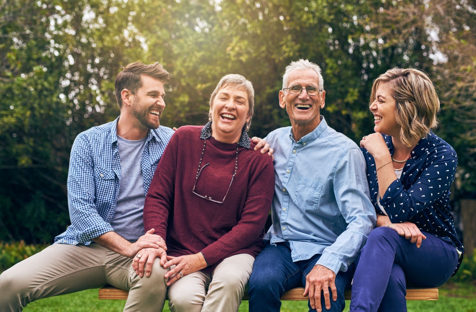 Four adults sitting on a wooden bench outdoors, laughing and enjoying a sunny day in a green park setting.
