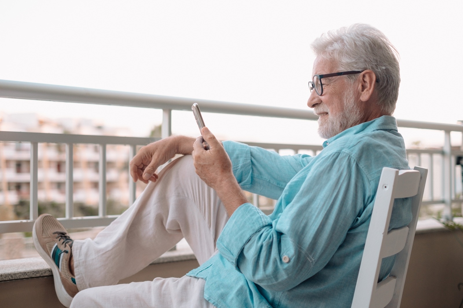 Older man with white hair and glasses sits on a balcony, looking at his smartphone.